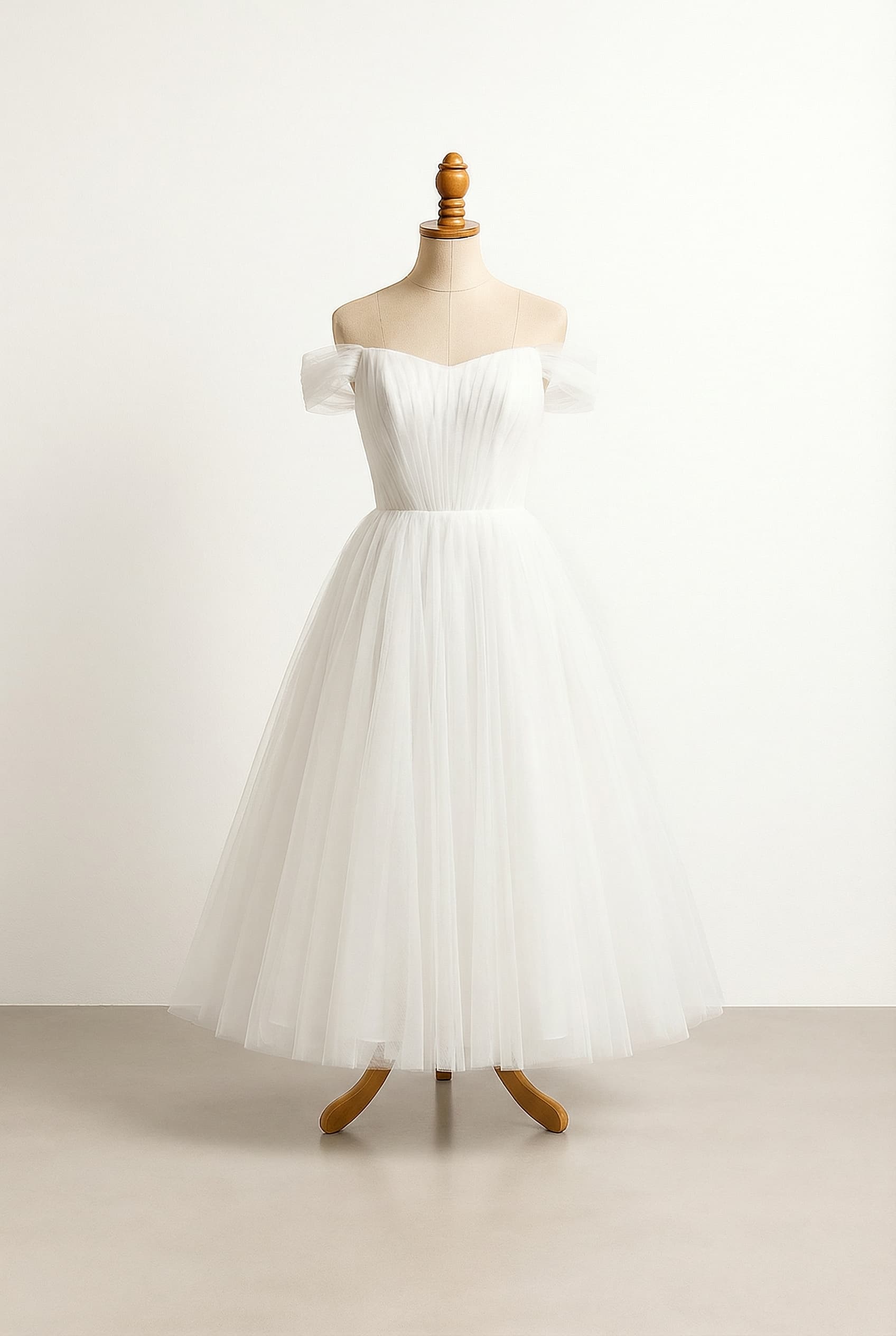 A romantic white A-line wedding dress with an off-the-shoulder, straight-across neckline. The bodice features delicate white lace embroidery, and the skirt flows gracefully in multiple layers of soft tulle, reaching a mid-calf length.