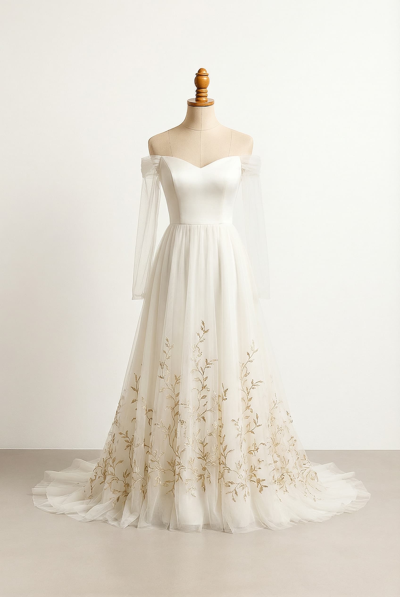 This wedding gown is an ethereal, romantic design with a flowing A-line silhouette, soft off-the-shoulder sweetheart neckline, and long, sheer tulle sleeves. The layered ivory tulle skirt falls into a soft, mist-like train. The clean, minimal bodice is free of lace or heavy embellishment. Delicate antique-gold vine embroidery is concentrated at the lower skirt and hem, growing upward organically and fading as it rises, ensuring the embellishment feels magical and restrained.