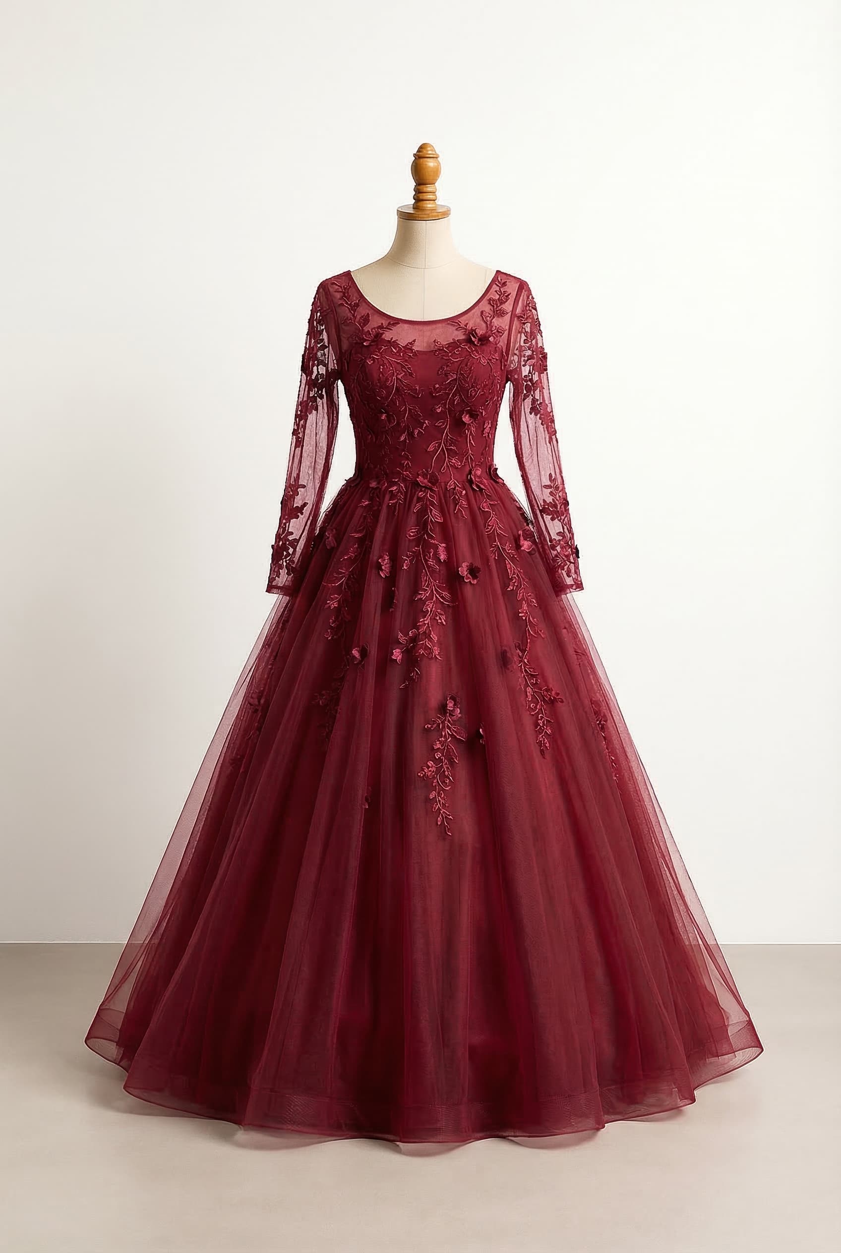A romantic burgundy ball gown crafted from layers of soft tulle creating a full flowing skirt that moves beautifully in the light The bodice features delicate embroidered lace vines and scattered floral appliqus that cascade gently into the skirt giving the dress an enchanting woodland feel The gown features a graceful halter neckline or a classic scoop neckline paired with sheer full-length lace sleeves embroidered with subtle floral details