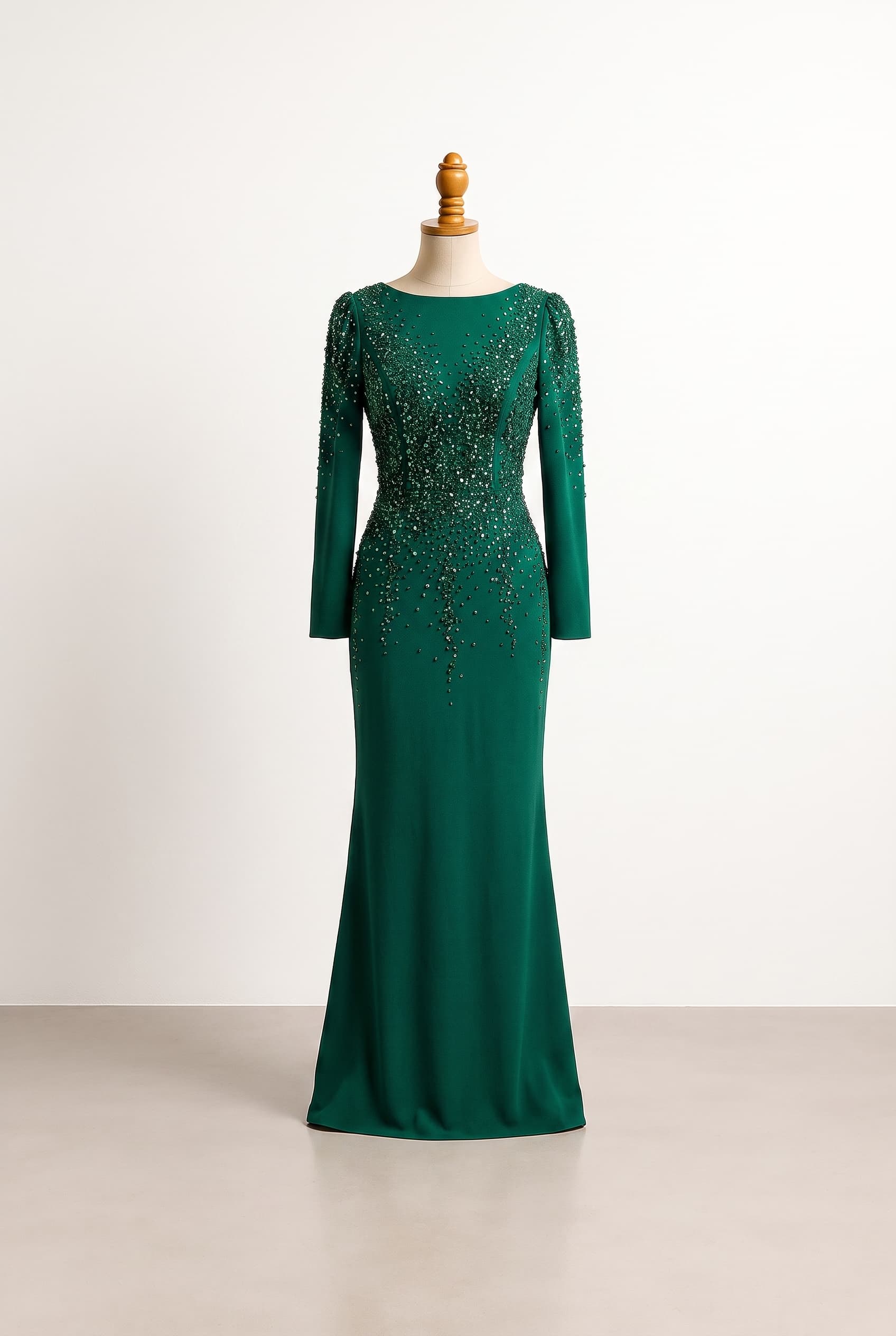 Hijab-friendly ultra-glam modest evening gown in a fitted column silhouette, deep emerald floor-length stretch crepe, full-length fitted sleeves, high bateau neckline, concentrated refined crystal and sequin embellishment across the bodice and shoulders tapering into a delicate scattered cascade down the skirt, high modest back with concealed zip